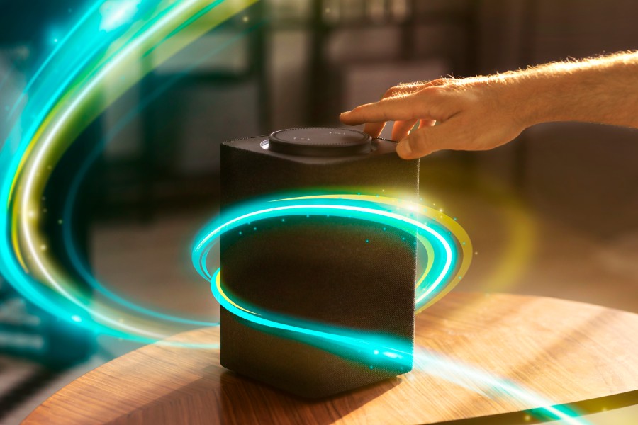 A hand reaches toward a black smart speaker on a wooden table, surrounded by glowing turquoise and green light trails.